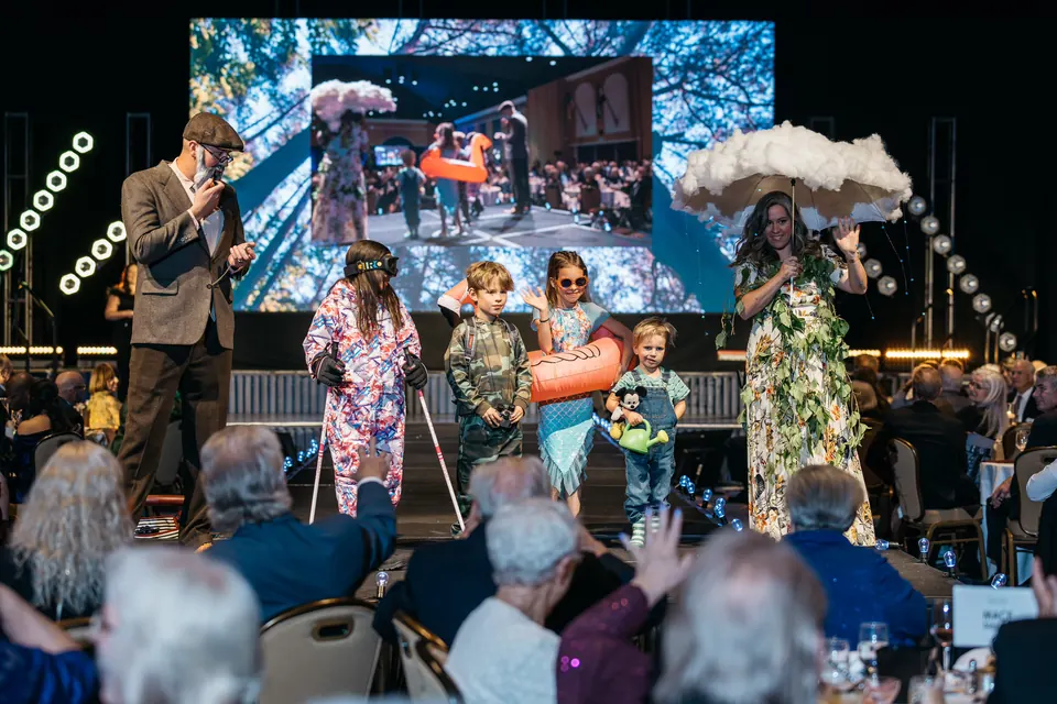 Models in costume walking the stage in front of a large video screen