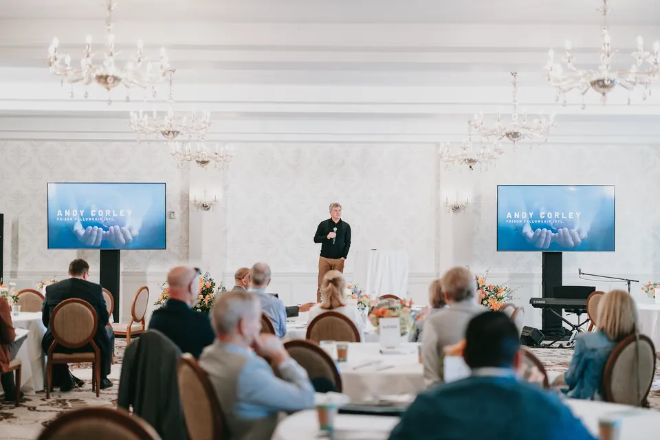 Presenter speaking to guests at round tables, with slides on two screens