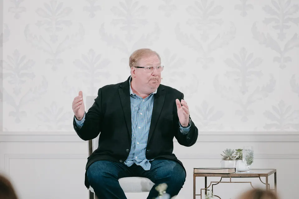 Speaker seated on stage in front of a white paneled wall